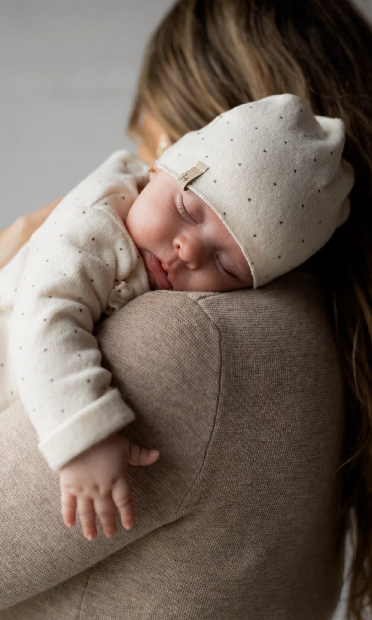 Newborn baby in a white outfit with small black dots, sleeping peacefully on a person's shoulder.