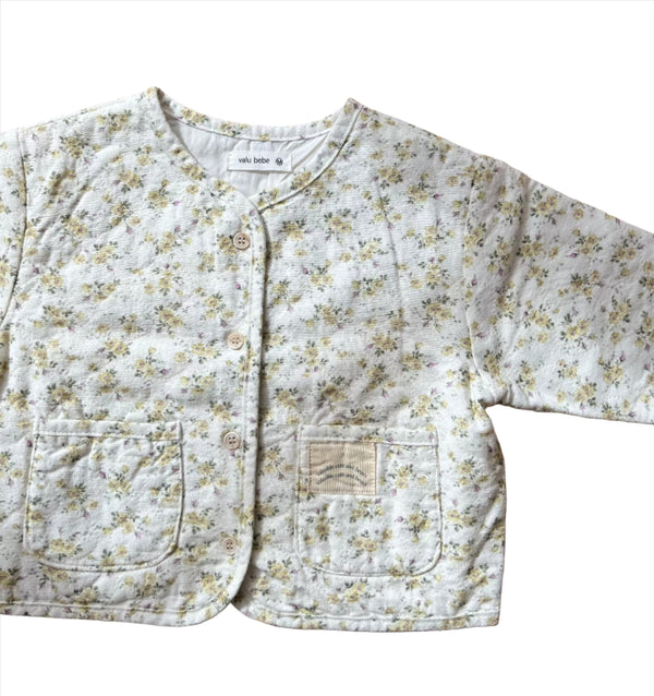 A lightweight quilted baby jacket with a floral pattern, featuring a v-neck and traditional wooden button closure, in earthy yellow and pink tones.