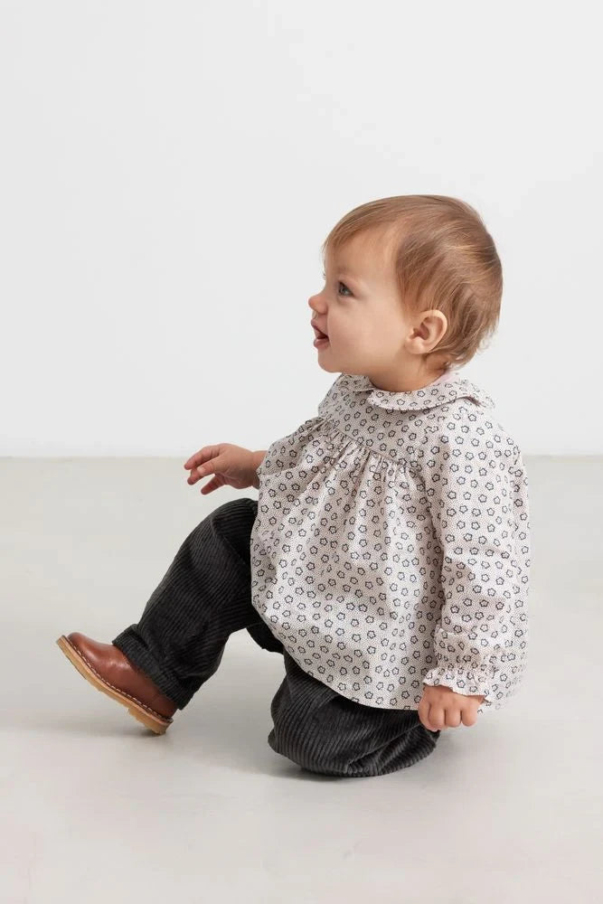 Baby sitting on a white surface wearing a patterned shirt and dark pants.